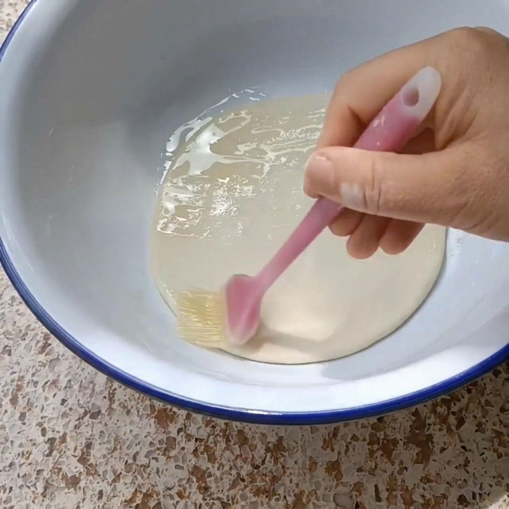 Una mano usando un pincel de silicona para cubrir un disco de masa redondo con aceite vegetal en un bol blanco.