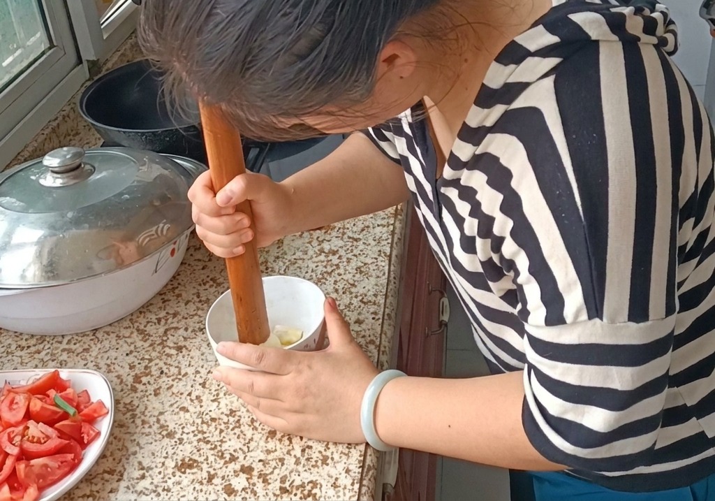 Una persona usando un mazo de madera para machacar dientes de ajo en una pasta suave en un bol blanco.
