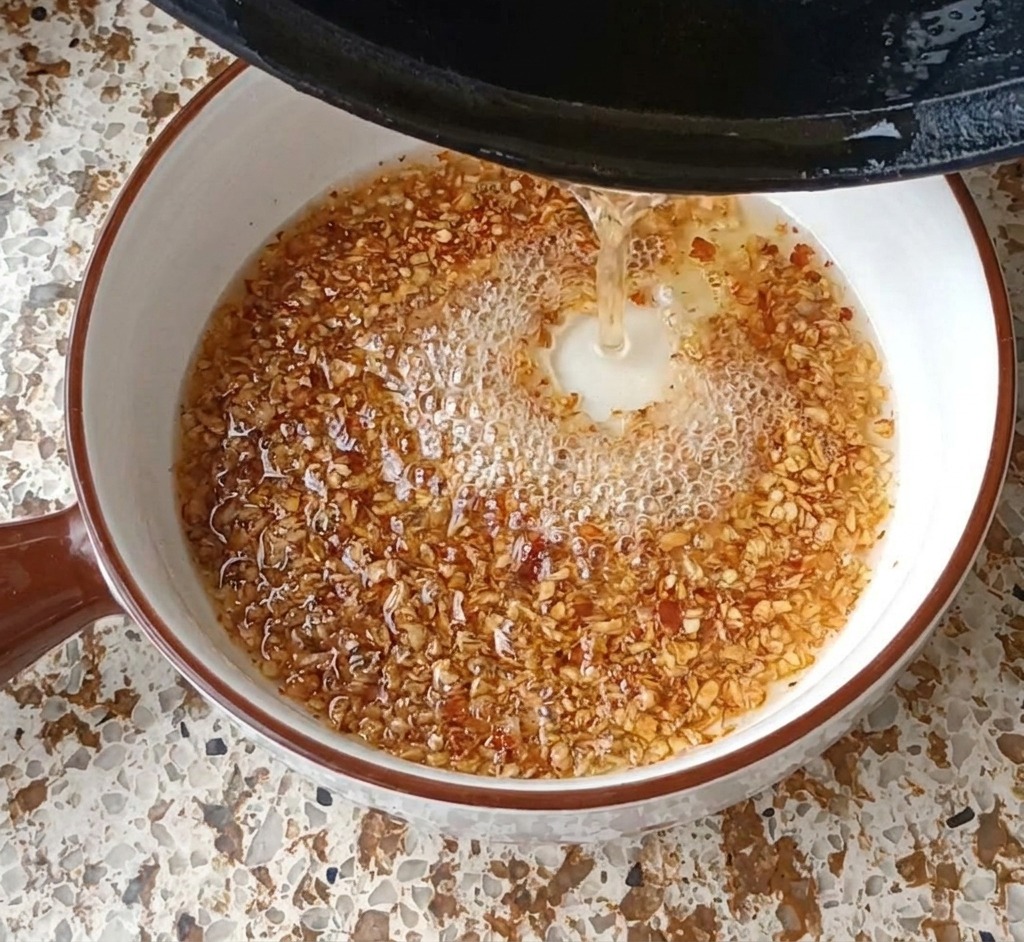 Aceite caliente siendo vertido desde una sartén negra en un bol blanco con ajo y chile, creando burbujas.