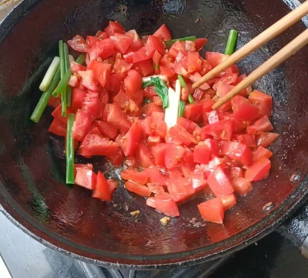 Tomates picados y trozos de cebolleta siendo salteados con palillos de madera en un wok de hierro fundido.
