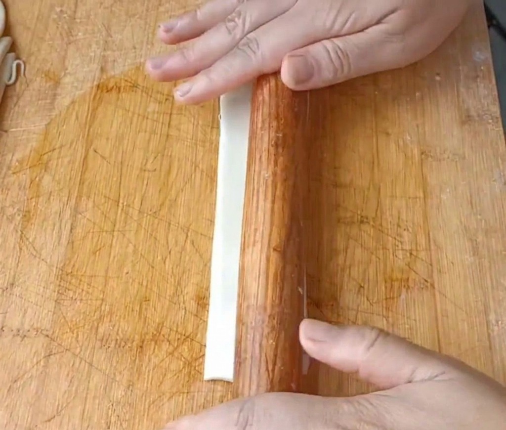Primer plano de manos usando un rodillo de madera para aplanar una tira larga de masa aceitada en una tabla de madera.