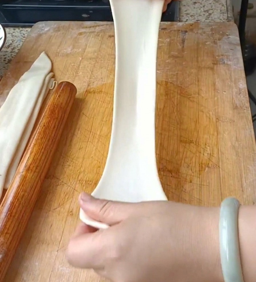 Dos manos estirando verticalmente una tira de masa ancha y traslúcida para formar un fideo largo.
