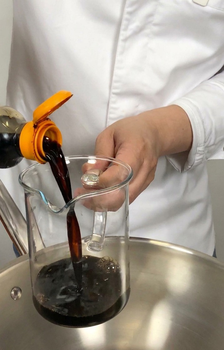 Salsa de soja oscura vertiéndose desde una botella en una jarra medidora sobre una cacerola de acero inoxidable.