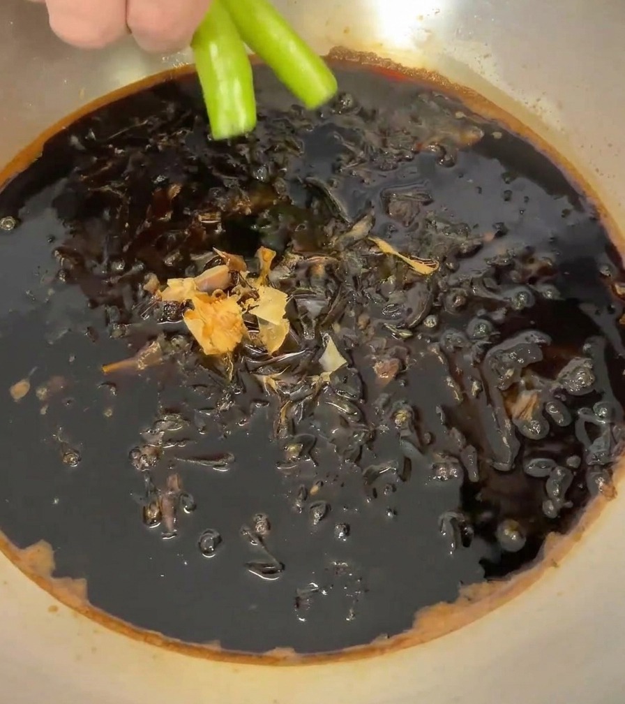 Una mano colocando dos chiles verdes brillantes en la cacerola llena de salsa oscura y copos de bonito.