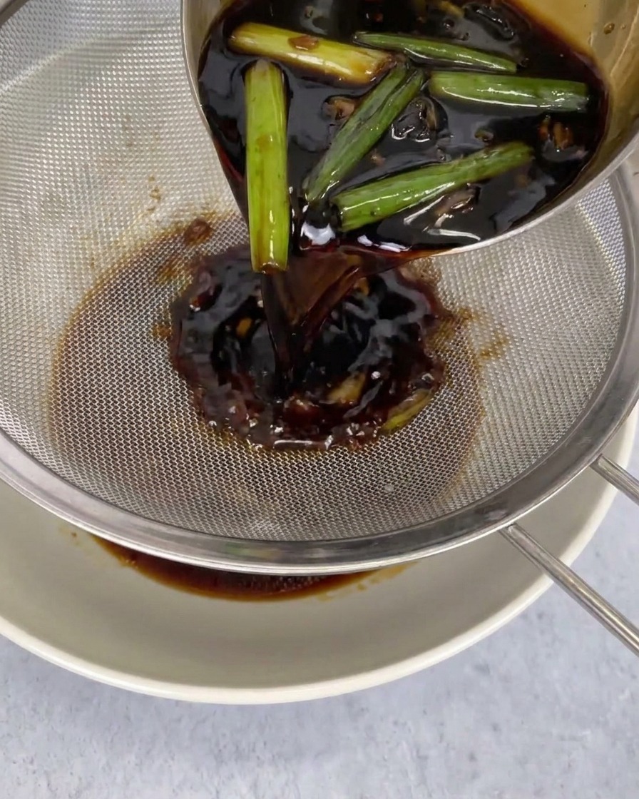 Salsa oscura vertiéndose desde un cazo a través de un colador metálico hacia un bol blanco, atrapando los chiles y otros ingredientes.