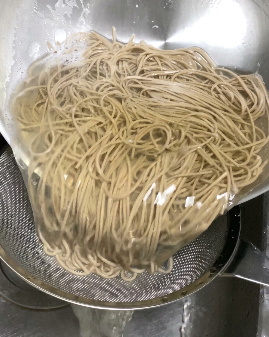 Fideos soba cocidos vertiéndose desde un bol grande a un colador metálico en el fregadero para escurrir el líquido.