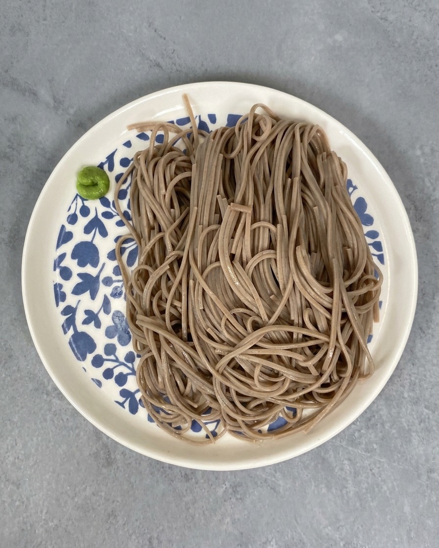 Una ración de fideos soba cocidos en un plato blanco con detalles florales azules, con una pequeña porción de pasta de wasabi al lado.