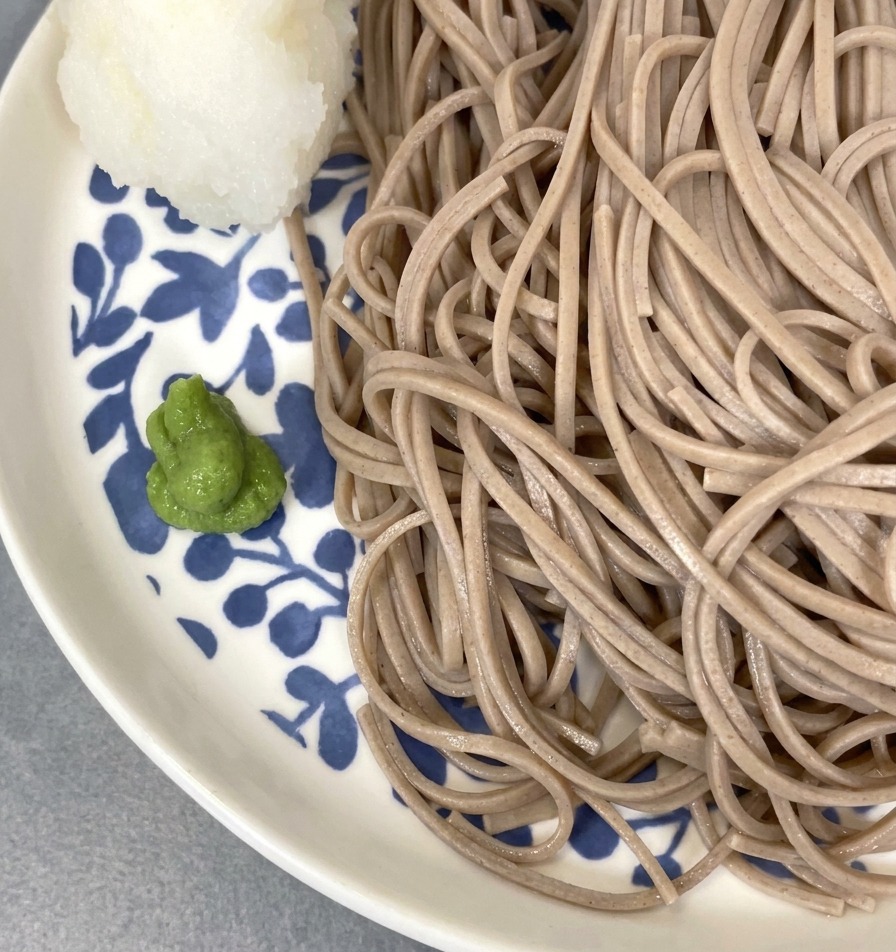 Primer plano de fideos soba fríos servidos en un plato floral, decorados con wasabi y una bola de rábano rallado moldeada.