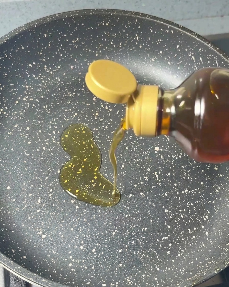 Aceite de sésamo vertiéndose desde una botella en una sartén oscura moteada en un movimiento circular constante.