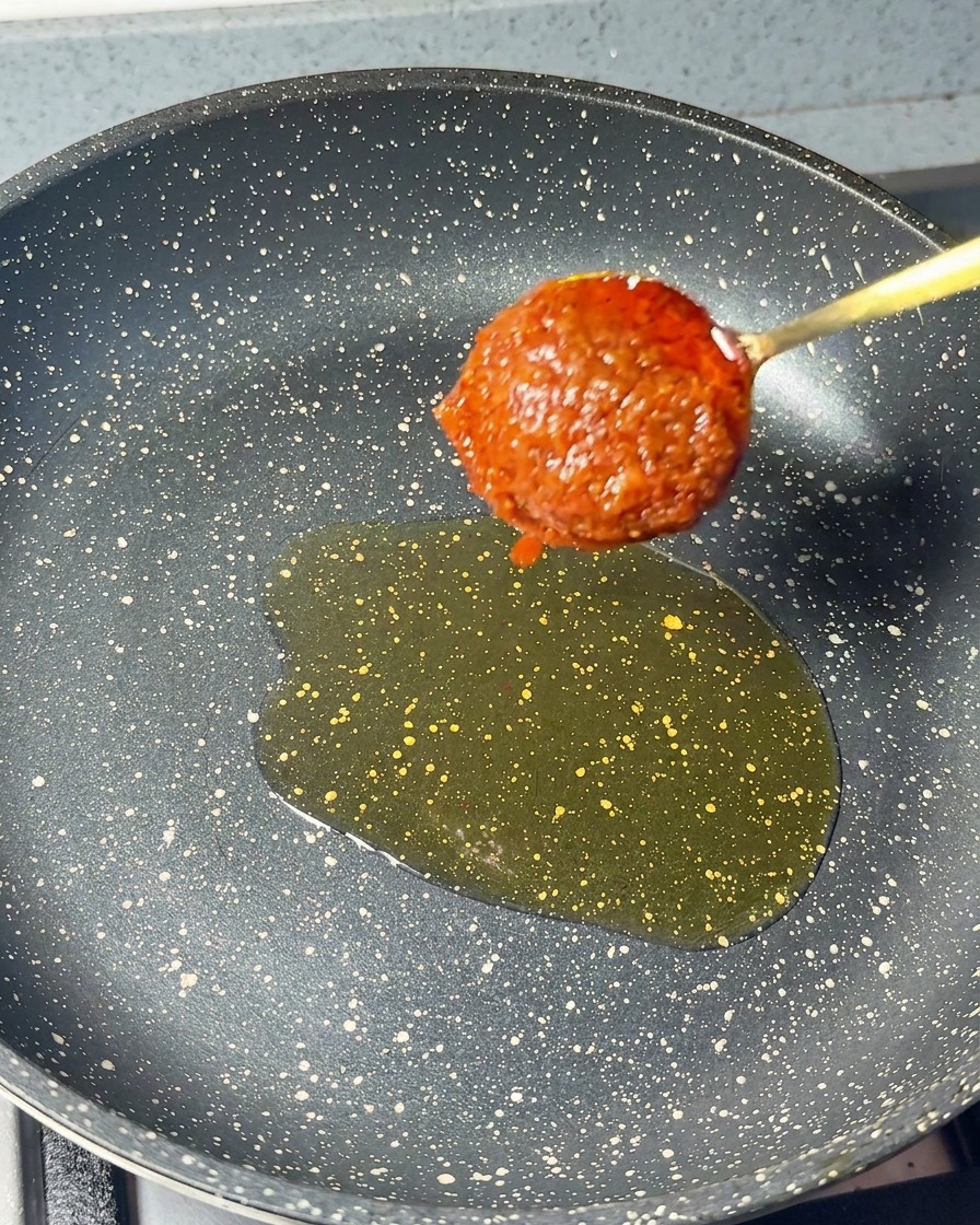 Una cucharada de pasta de camarones roja espesa bajando hacia una sartén con un poco de aceite de sésamo.