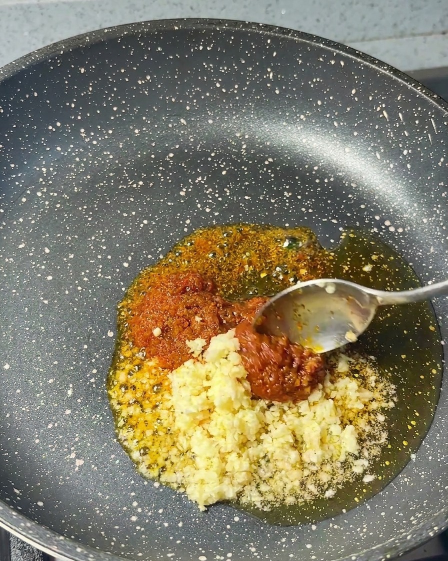 Ajo picado cayendo desde una cuchara a una sartén que ya contiene pasta de camarones y aceite de sésamo.
