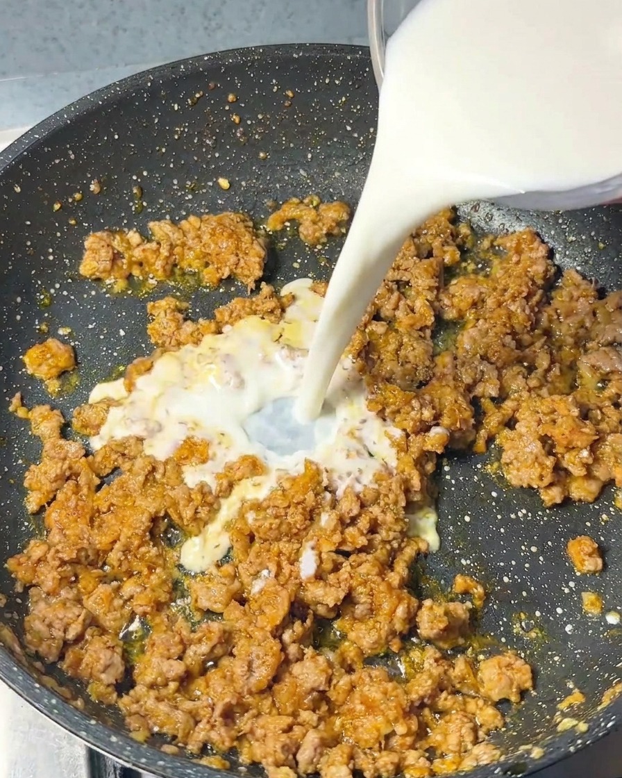 Un chorro de leche blanca vertiéndose desde un recipiente de vidrio en una sartén llena de cerdo cocido y especias.