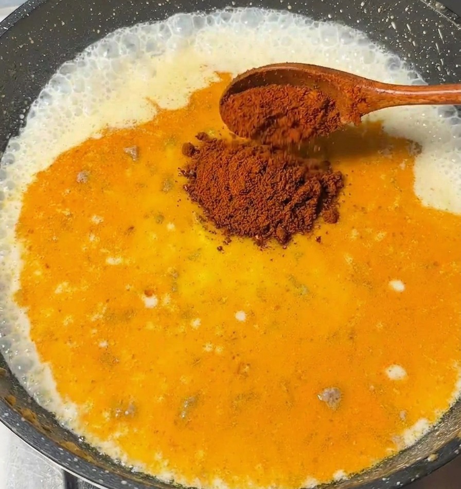 Una cuchara de madera añadiendo trozos de roux de curry en una salsa a base de leche que burbujea en una sartén oscura.