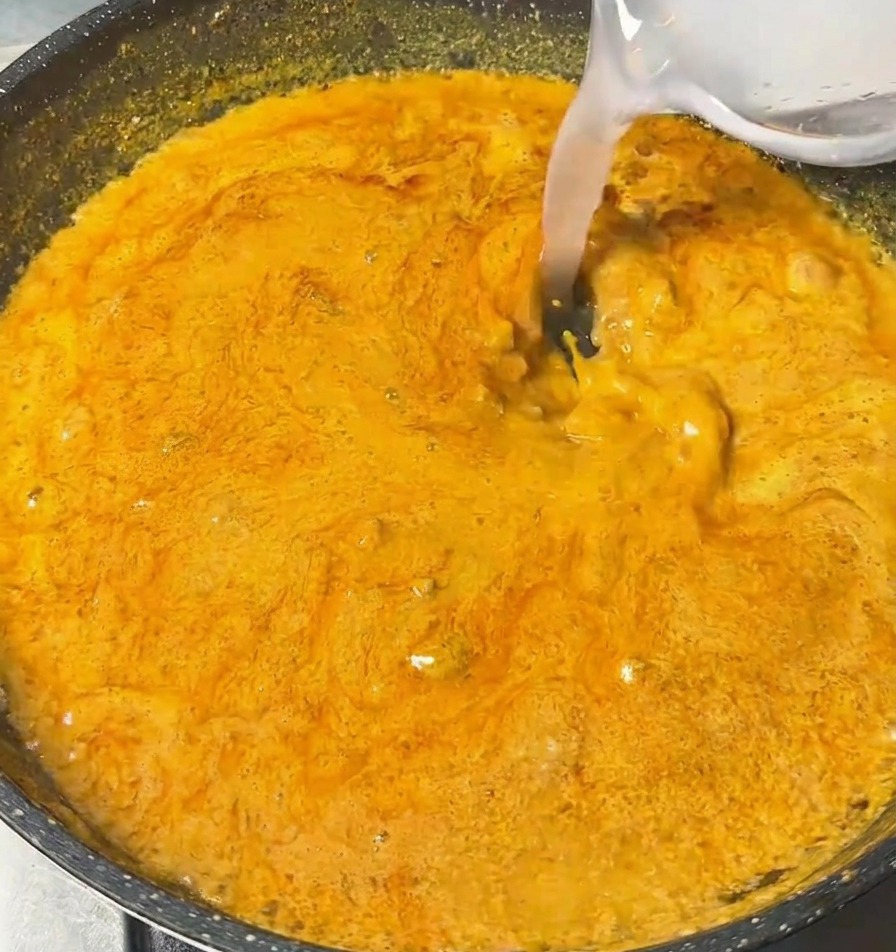 Un líquido claro vertiéndose desde un bol pequeño en una sartén con salsa de curry naranja espesa y burbujeante.