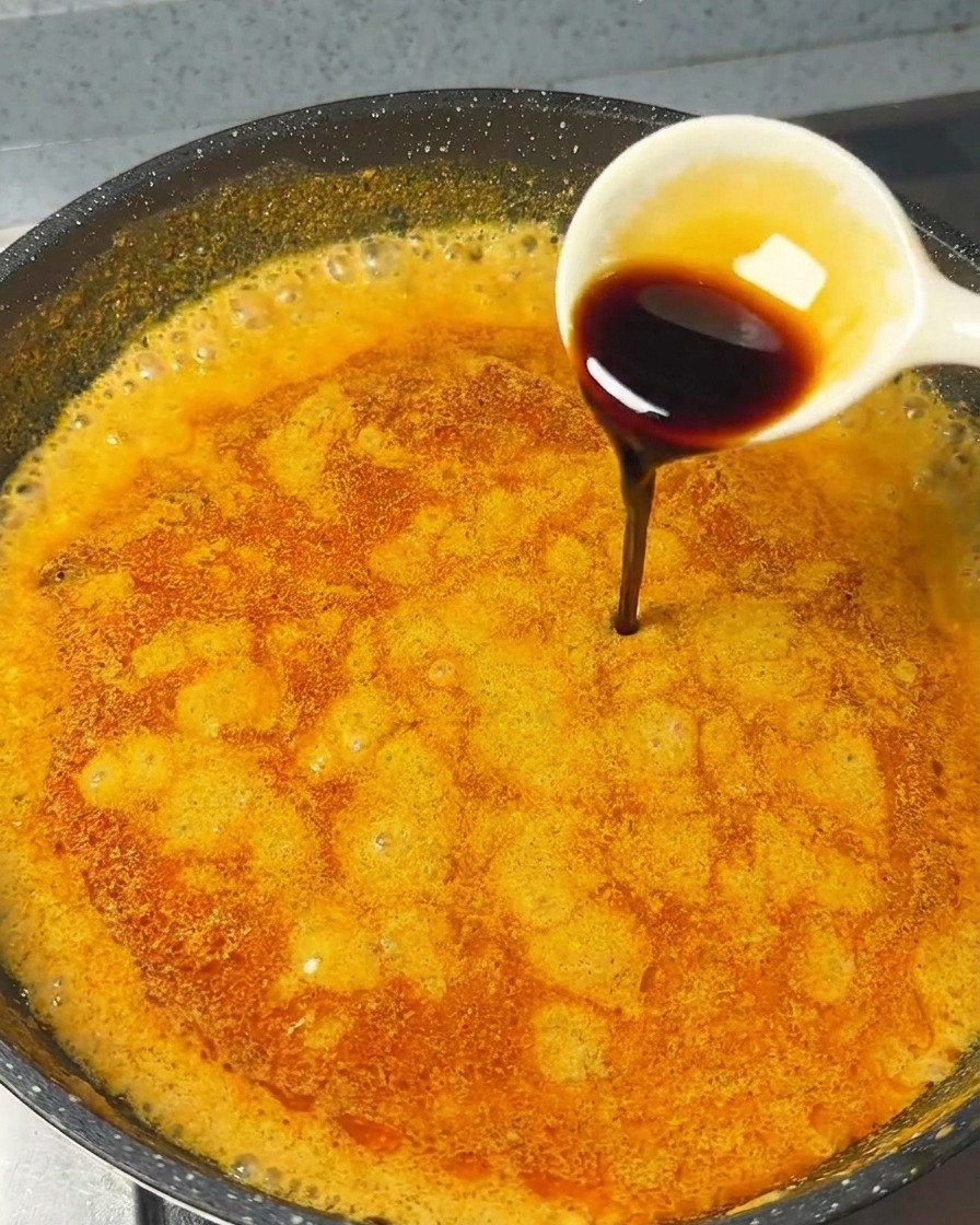 Una cuchara blanca vertiendo salsa de soja oscura en una salsa de curry naranja que burbujea.