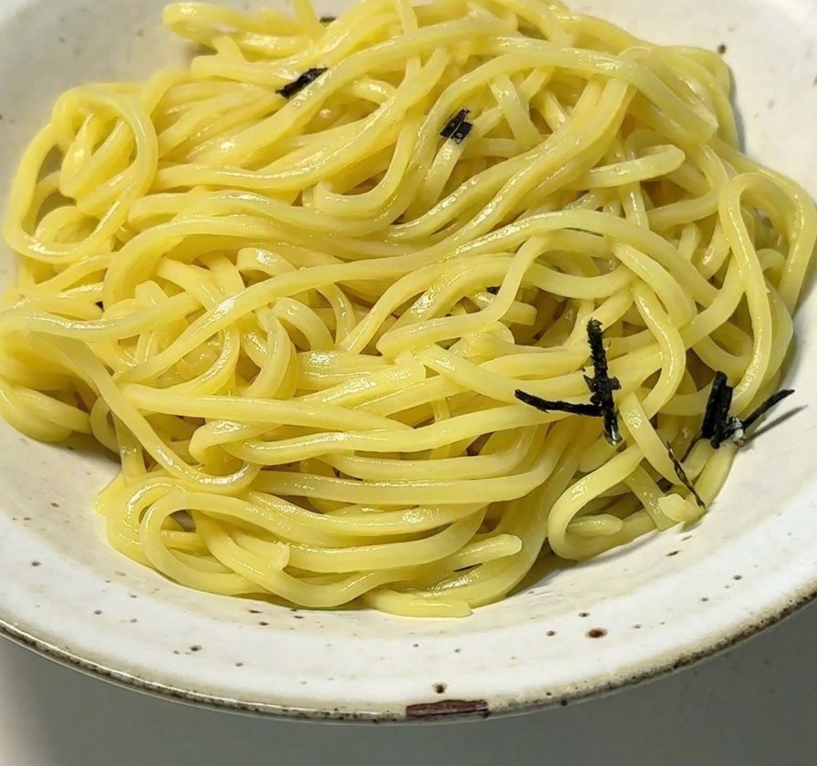 Un bol de cerámica blanca lleno de fideos ramen amarillos coronados con tiras de alga nori seca negra.