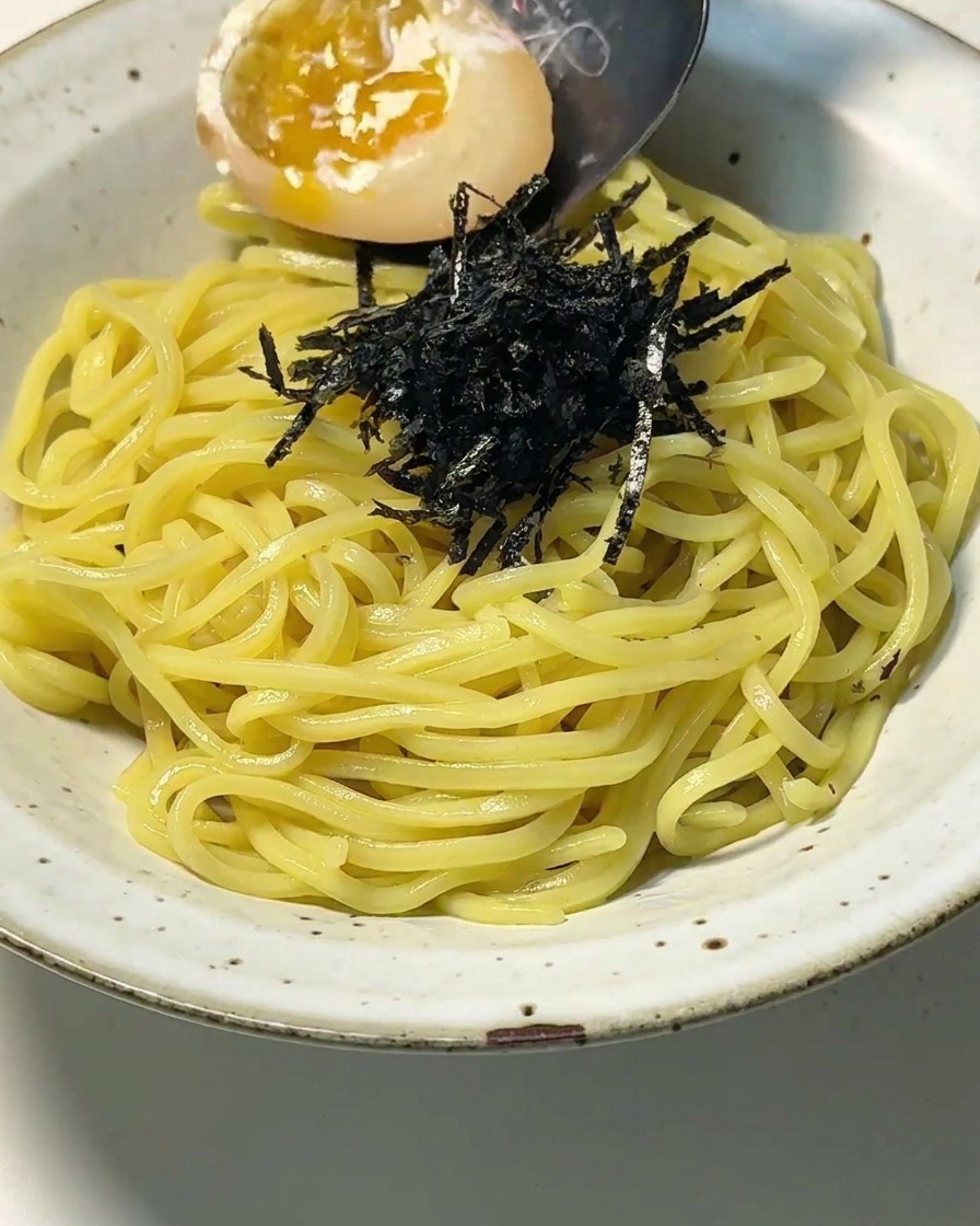 Vista de cerca de un bol con fideos y nori mientras una cuchara coloca un huevo partido por la mitad con una yema líquida y brillante.