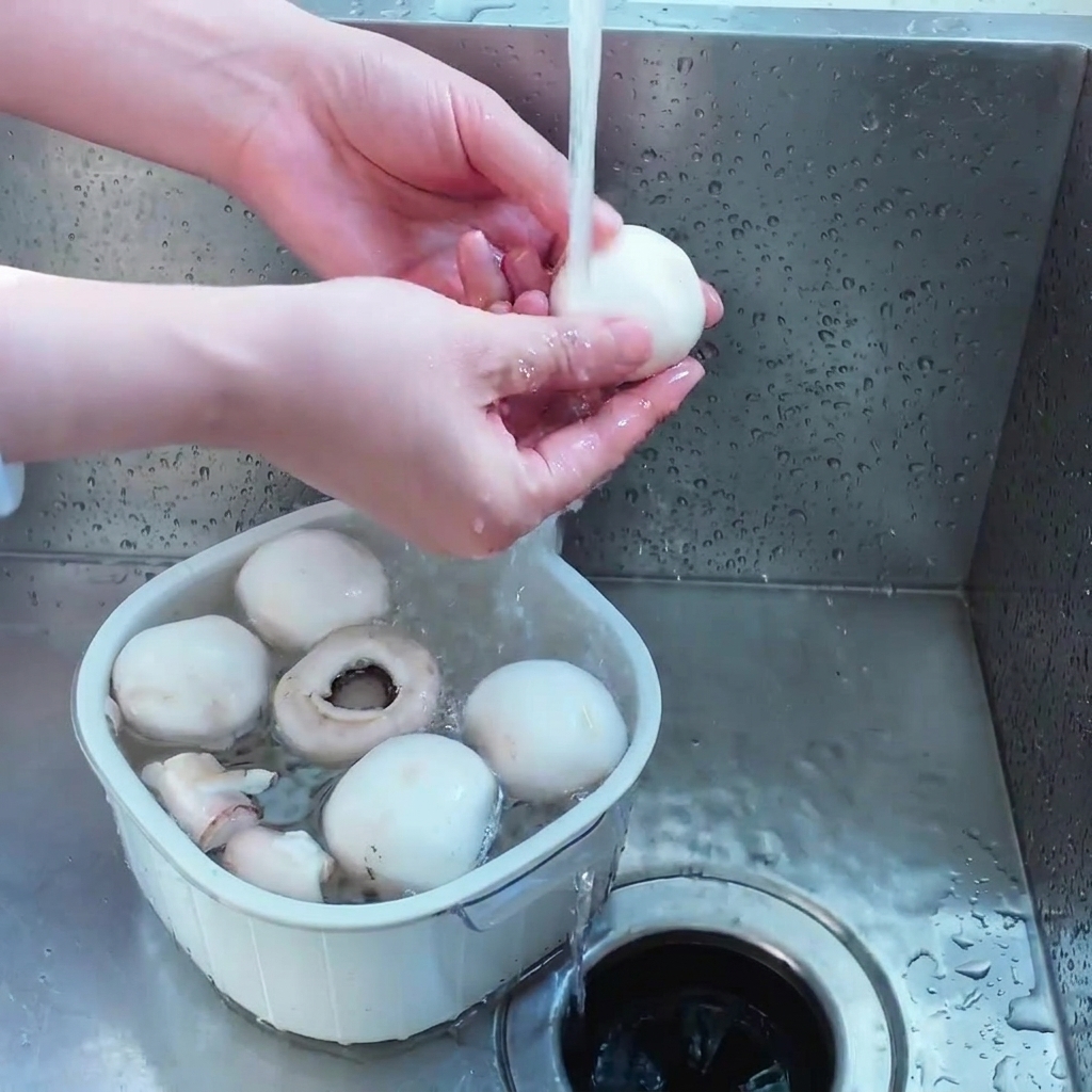 Una persona lavando varios champiñones blancos enteros en un colador de plástico blanco bajo el chorro de agua del grifo.