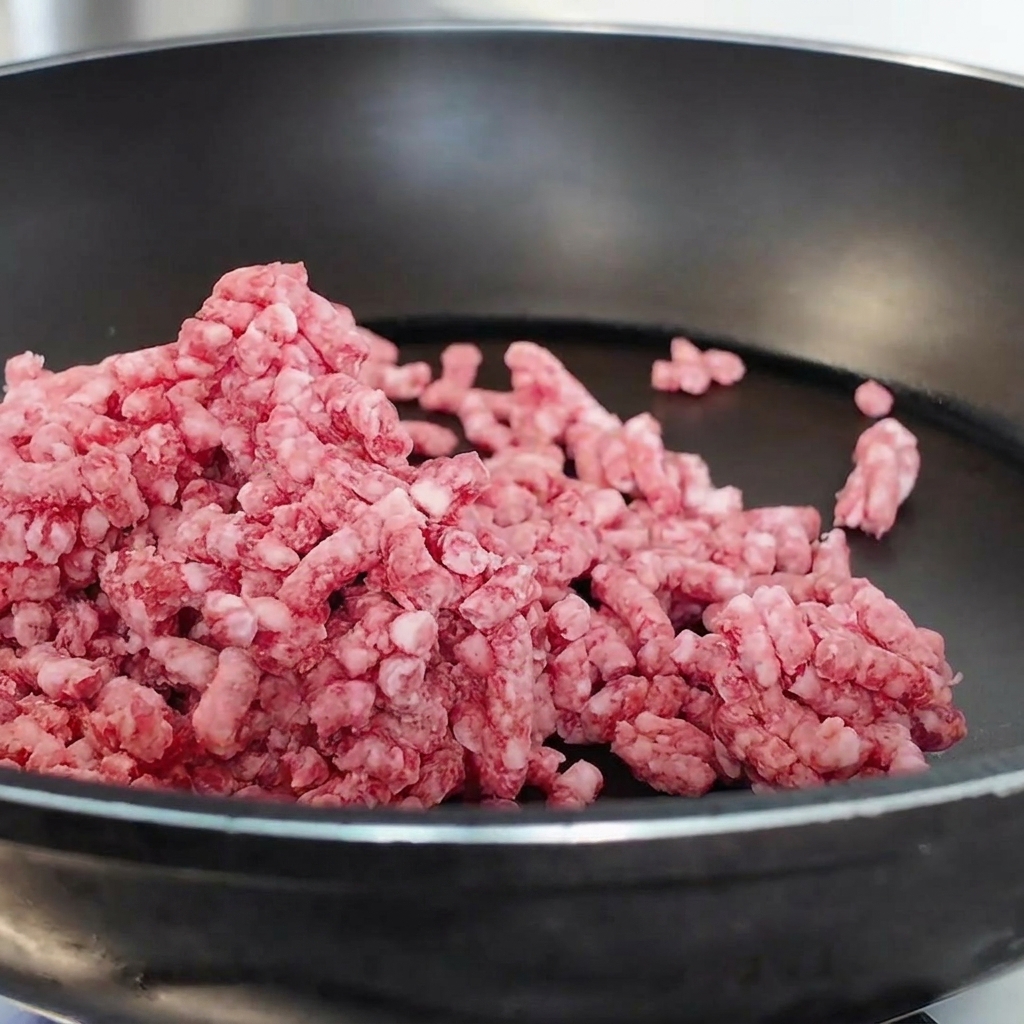 Carne picada de ternera cruda siendo desmenuzada y salteada en una sartén antiadherente negra.
