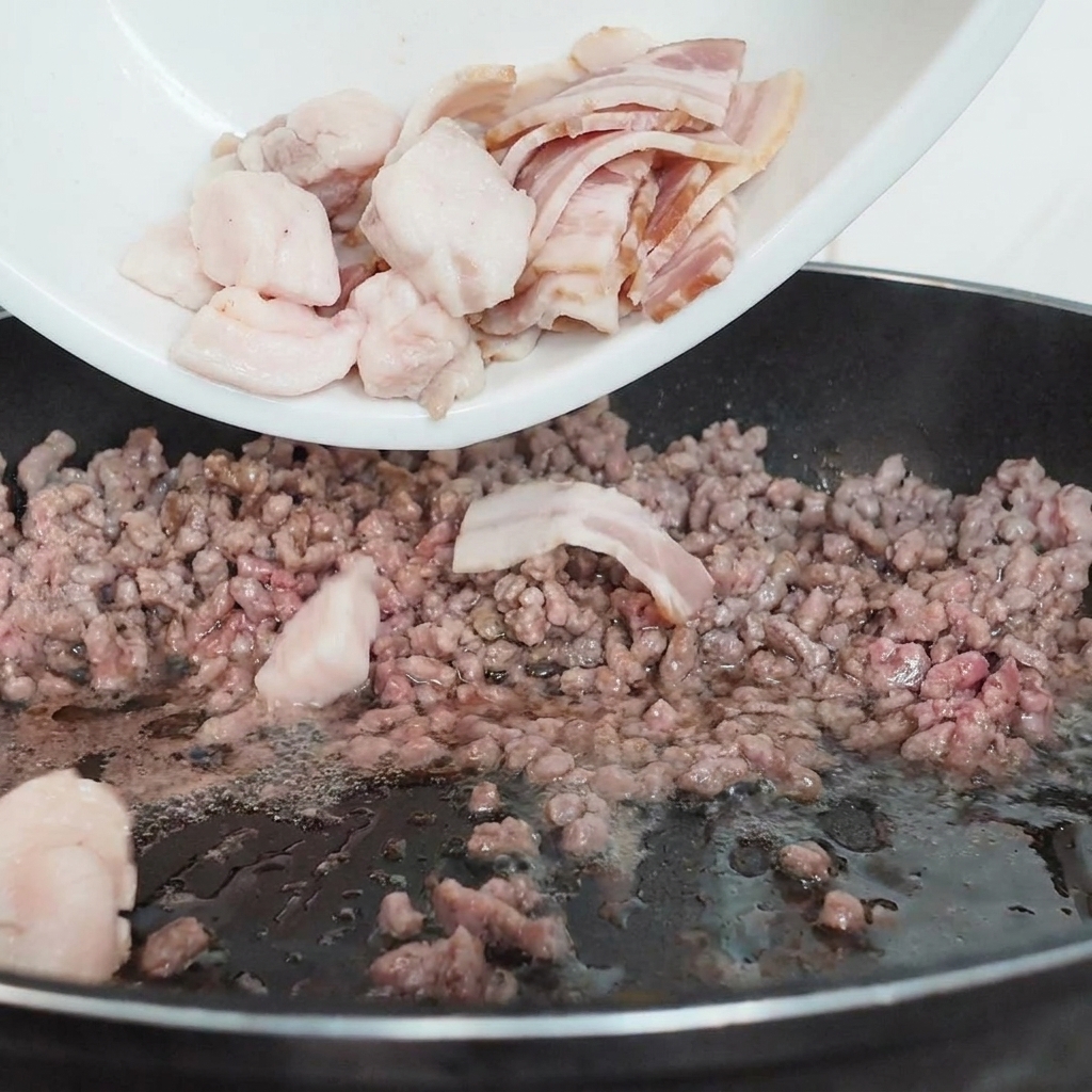 Rodajas de champiñón blanco vertiéndose desde un plato blanco a una sartén negra que contiene carne picada parcialmente cocida.