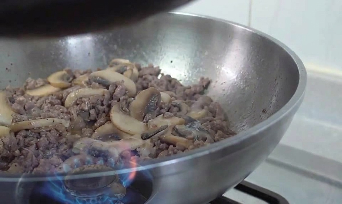 Un wok de acero inoxidable que contiene una mezcla de carne picada dorada y rodajas de champiñón salteadas sobre una llama de gas.