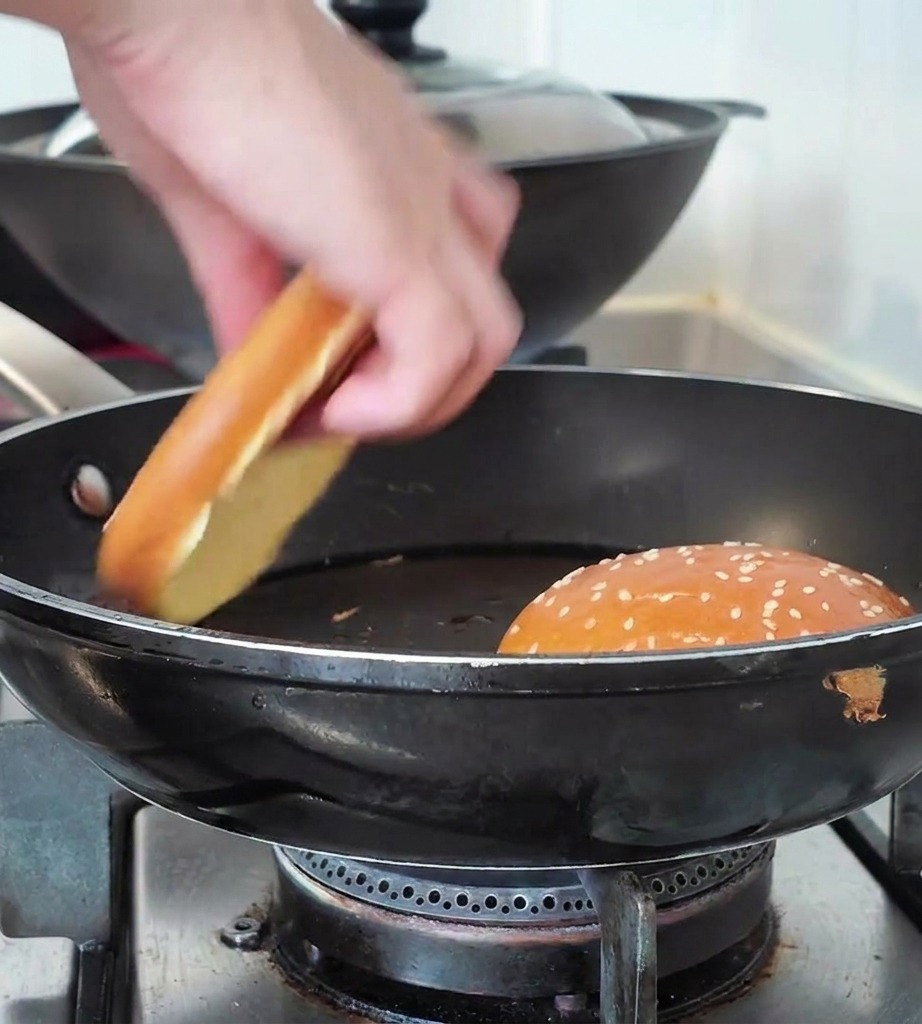 Una mano colocando la mitad de un pan de hamburguesa con sésamo en una sartén negra junto a otro pan tostándose.