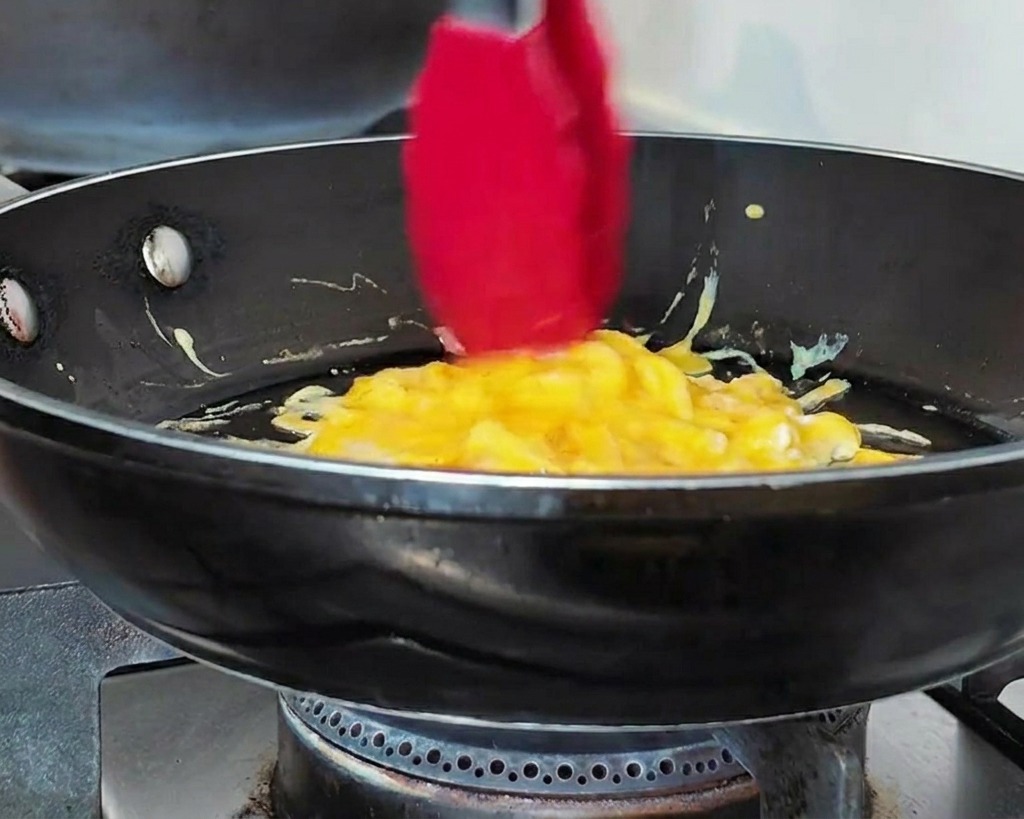 Pinzas rojas juntando huevos revueltos tiernos en forma circular en una sartén negra.
