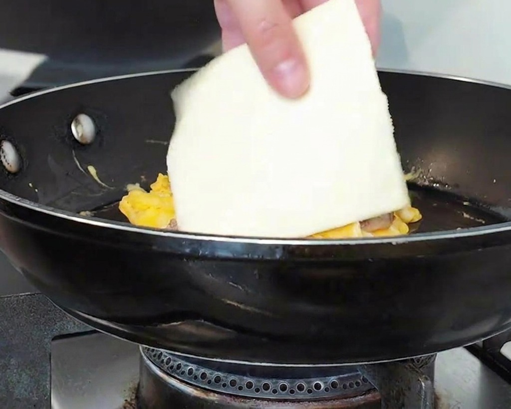 Una mano colocando una rodaja cuadrada de queso blanco sobre una montaña de carne y huevos en una sartén.