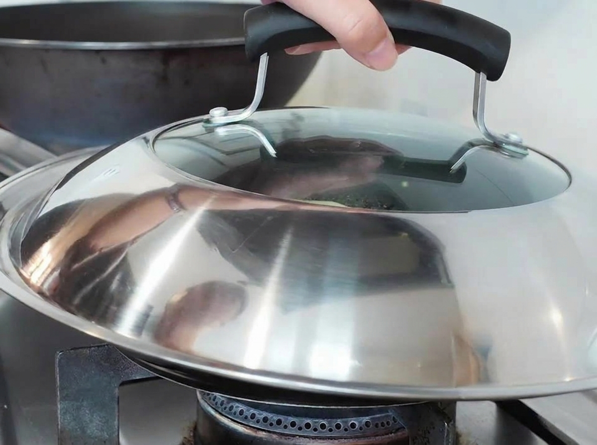Una mano colocando una tapa grande de metal y vidrio sobre una sartén en una estufa de gas.