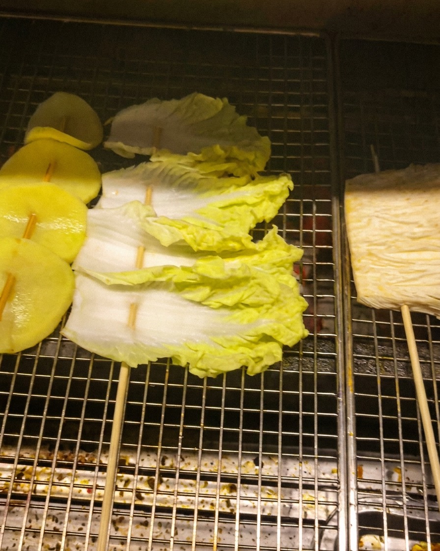 Rodajas de patata, hojas de col y setas enoki insertadas en brochetas sobre una rejilla de metal.