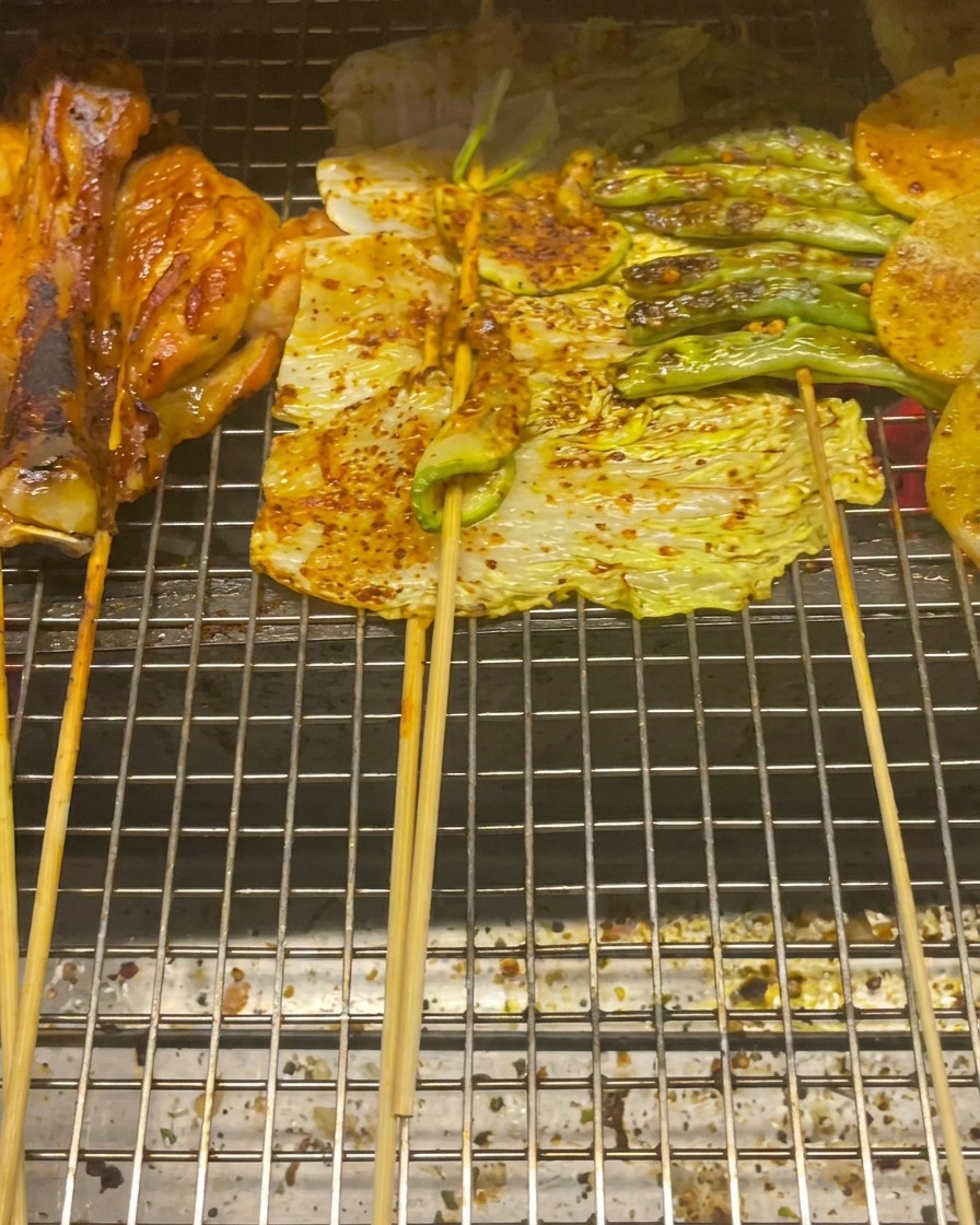 Contramuslos de pollo, col, calabacín, judías verdes y rodajas de patata asándose en una rejilla.