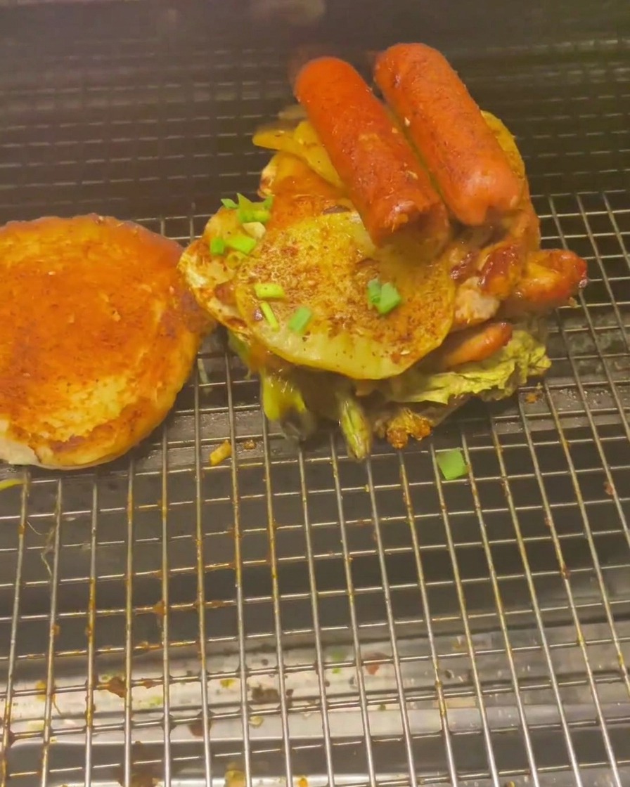 Una pila alta de carnes y vegetales a la parrilla, incluyendo salchichas y patatas, sobre el pan de hamburguesa.