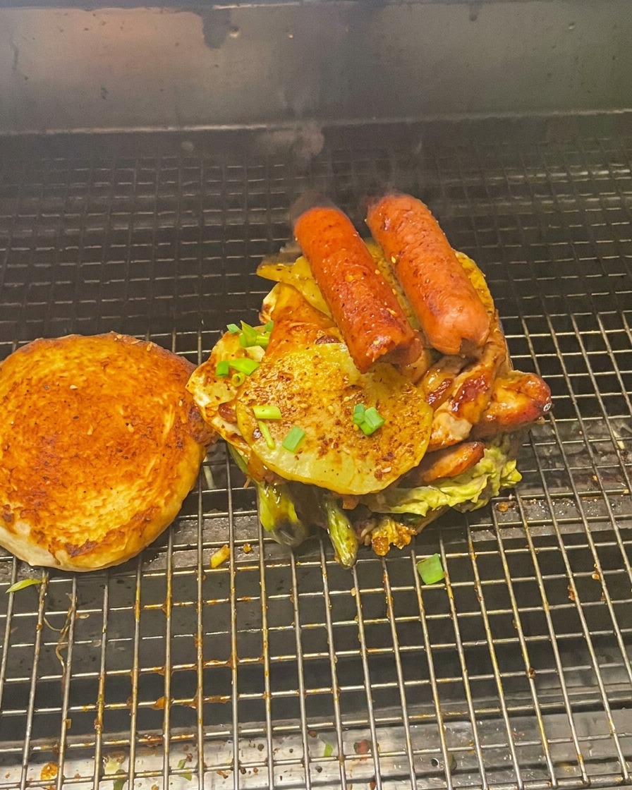 Pila gigante de carnes y vegetales en el pan inferior, con el pan superior tostado al lado en la parrilla.