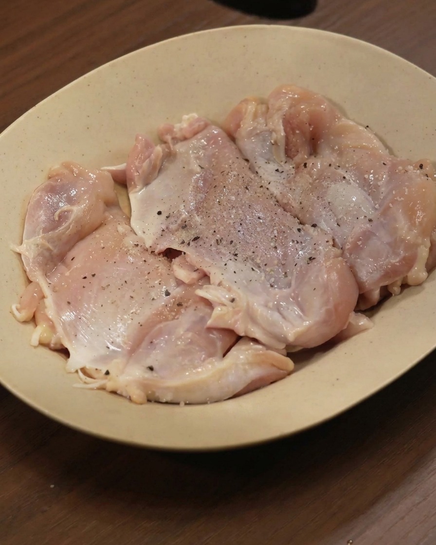 Contramuslos de pollo crudos en un plato beige, espolvoreados con pimienta negra y diversas especias molidas.