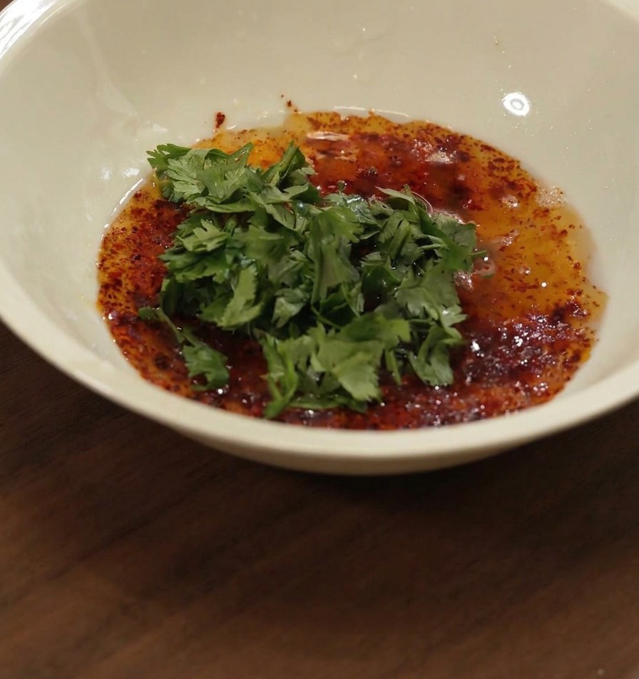 Miel vertiéndose en un bol blanco que contiene una mezcla de aceite rojo, especias y cilantro verde picado.