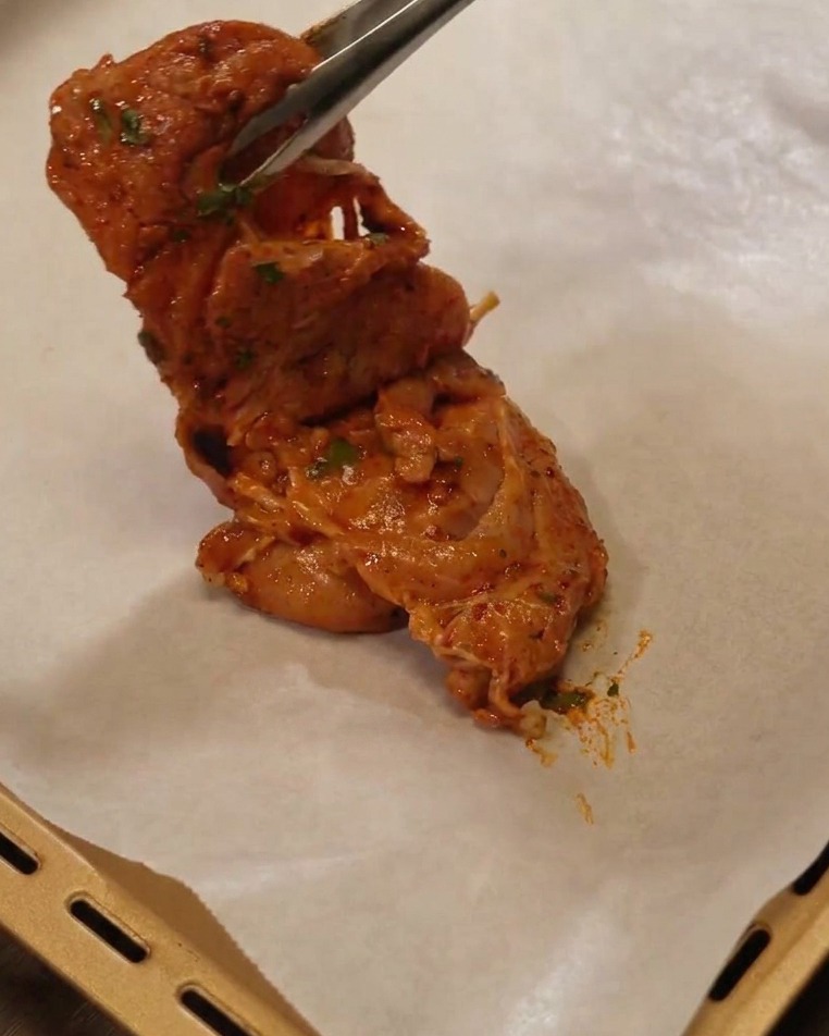 Pinzas de metal levantando un contramuslo de pollo marinado y colocándolo en una bandeja de horno con papel vegetal.