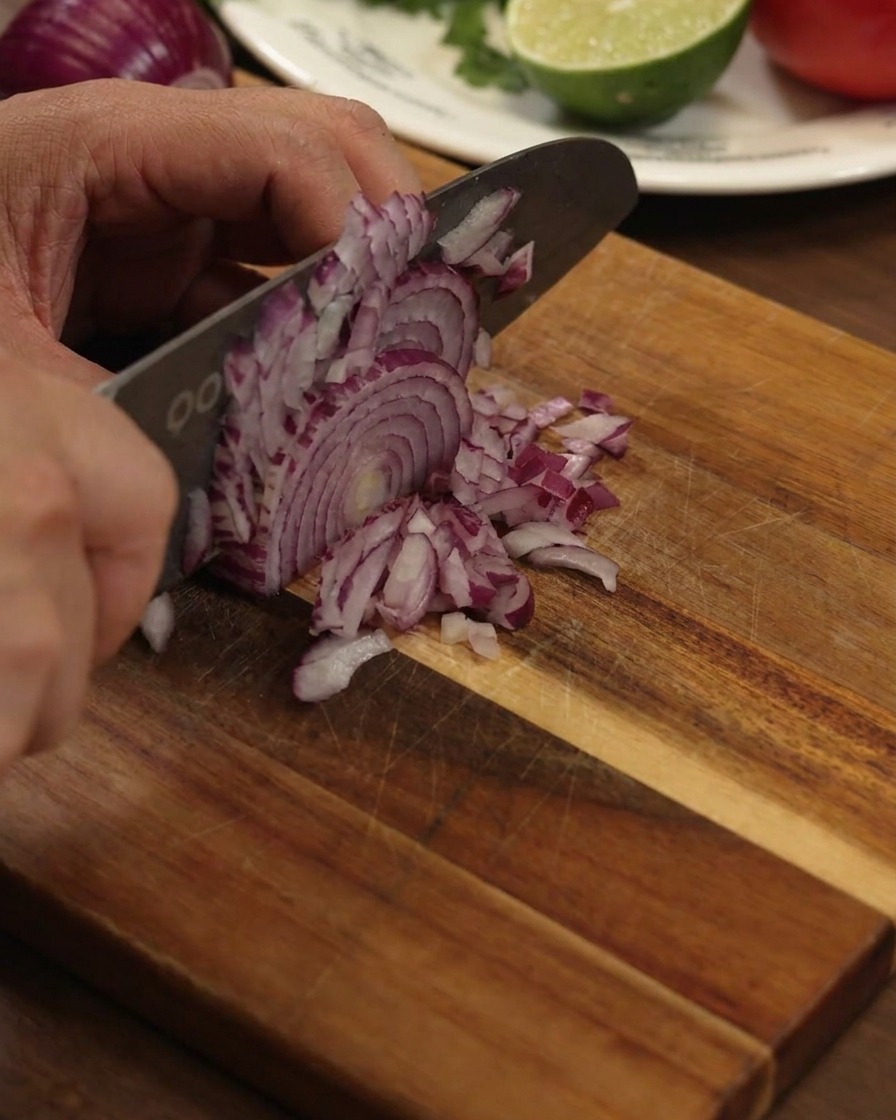 Una mano usando un cuchillo de cocina grande para picar finamente una cebolla roja sobre una tabla de madera.