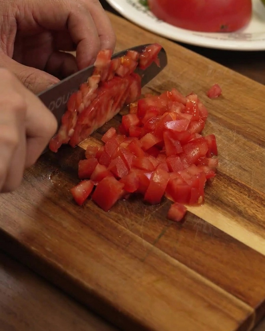 Cortando un tomate rojo maduro en dados pequeños y uniformes sobre una tabla de madera con un cuchillo de chef.
