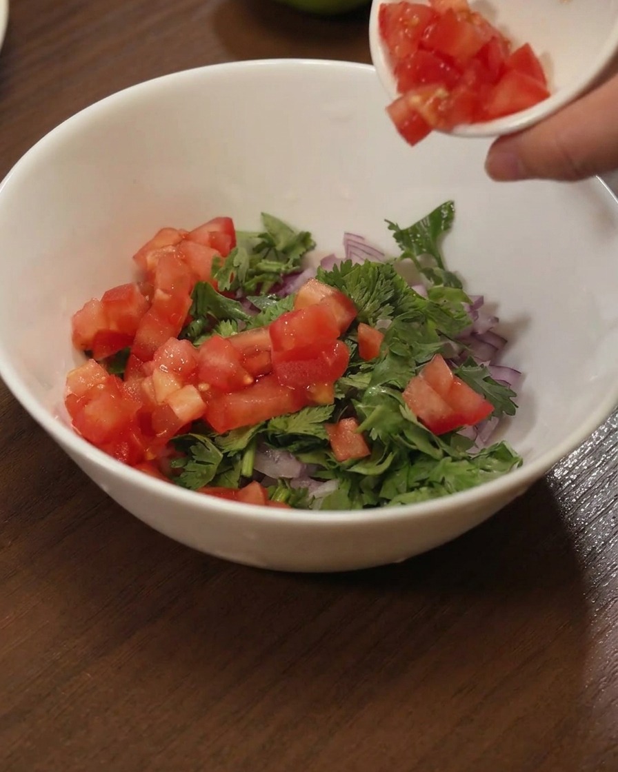 Dados de tomate rojo vertiéndose desde un plato pequeño a un bol blanco con cilantro y cebolla roja.