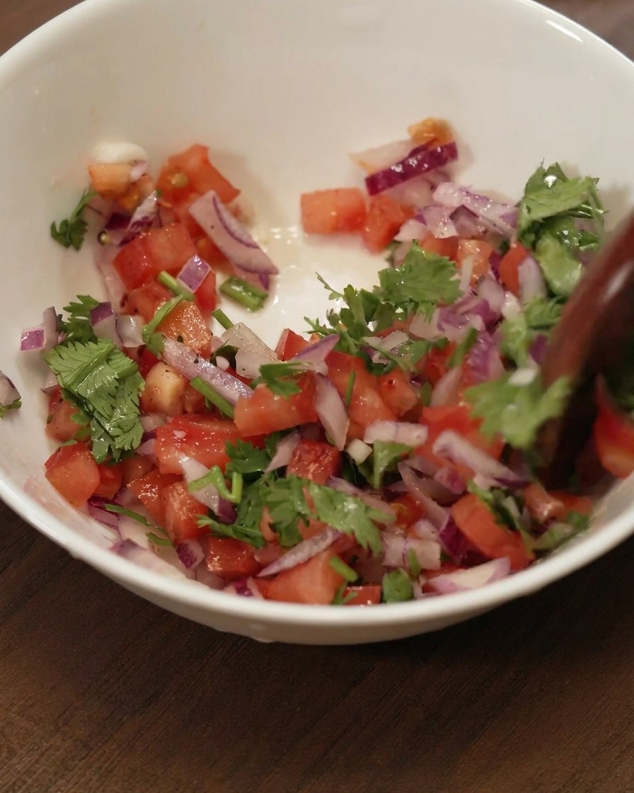 Una cuchara de madera removiendo una mezcla colorida de tomate, cebolla roja y cilantro en un bol cerámico blanco.