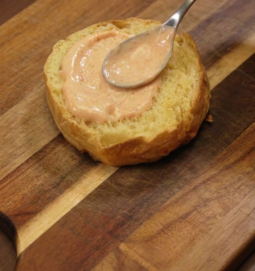Una cuchara de plata extendiendo una capa generosa de salsa naranja en la cara tostada de un pan brioche.