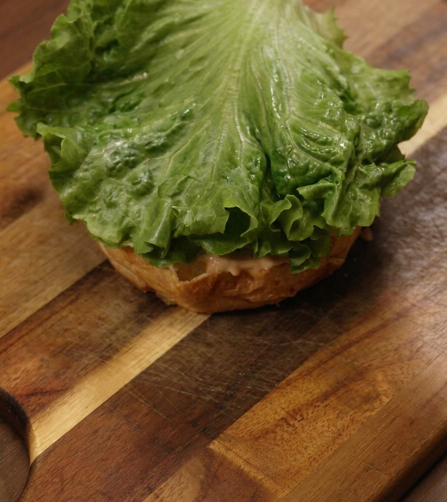 Una hoja de lechuga verde vibrante sobre un pan de hamburguesa con salsa en una tabla de madera.