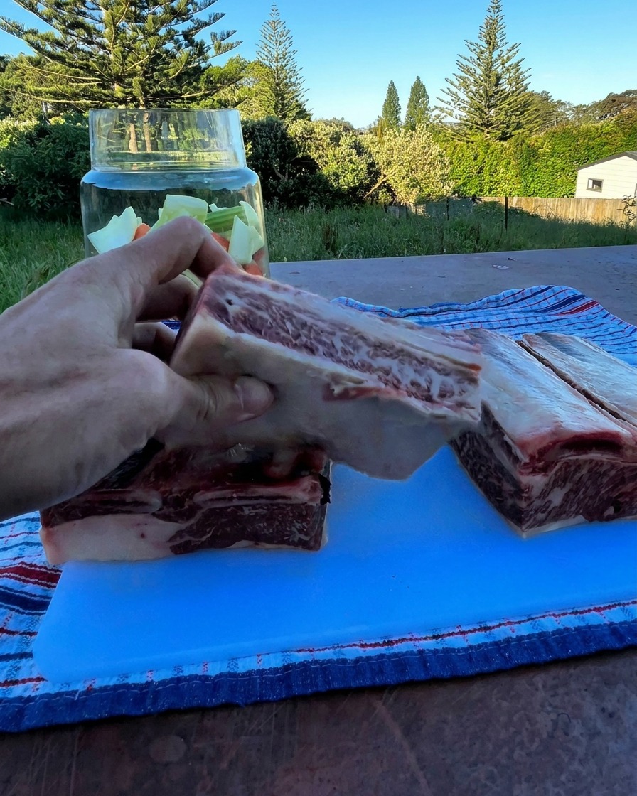Una mano sosteniendo una gruesa costilla de res cruda sobre un frasco de vidrio con vegetales picados.