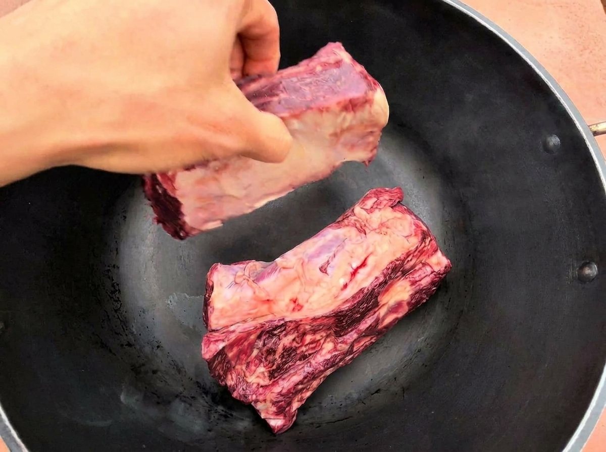 Una mano colocando dos grandes costillas de res crudas en una sartén negra caliente.