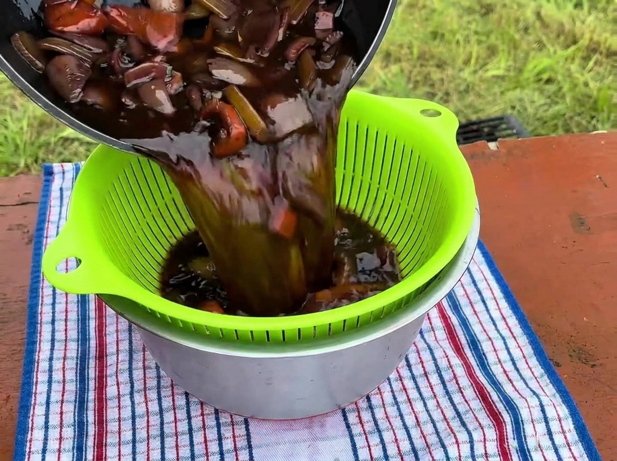 Líquido de braseado oscuro junto con vegetales cocidos vertiéndose de una olla a un colador verde sobre un tazón de acero inoxidable.