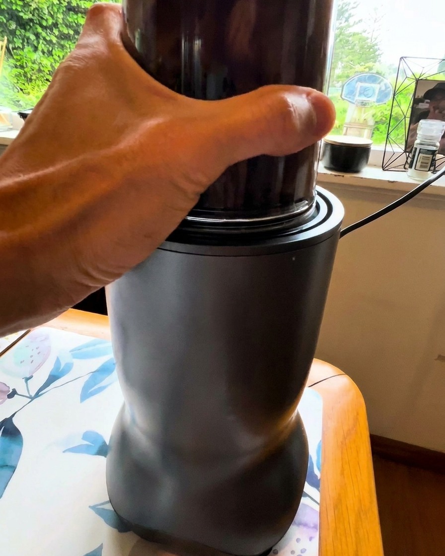 Una mano sosteniendo un vaso de licuadora pequeño mientras procesa una mezcla oscura de vegetales.