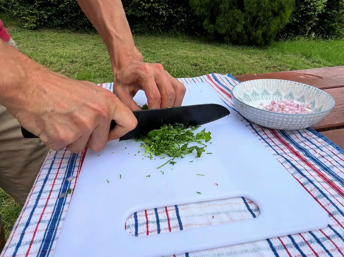 Manos de una persona usando un cuchillo negro para picar finamente perejil verde fresco en una tabla de cortar blanca junto a un tazón con chalotas picadas.
