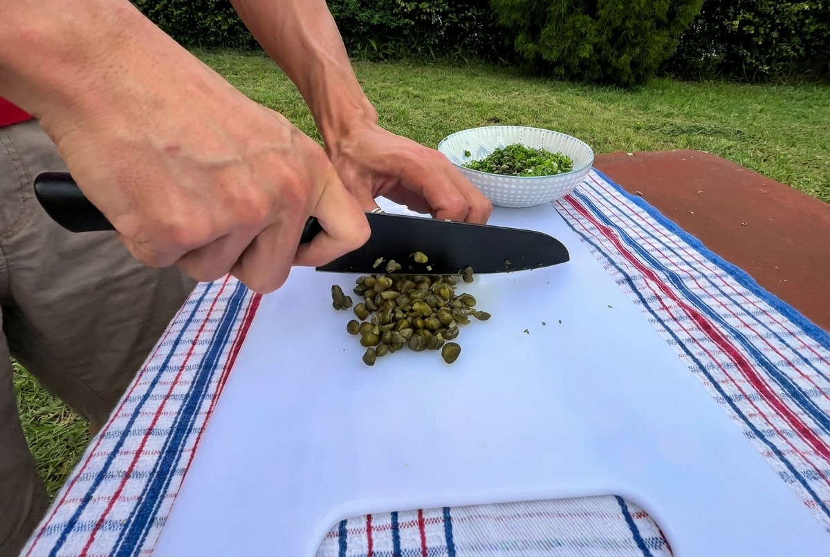 Un primer plano de una persona usando un cuchillo de chef negro para picar finamente alcaparras verdes en una tabla de cortar blanca al aire libre.