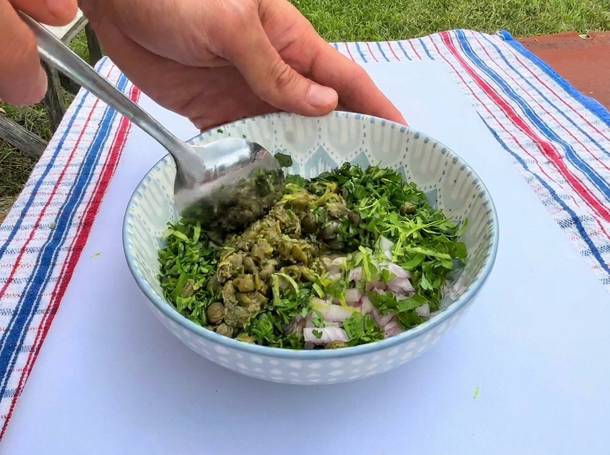 Mezclando una vibrante combinación de hierbas verdes picadas, alcaparras y chalotas finamente picadas con una cuchara de metal dentro de un tazón de cerámica decorado.