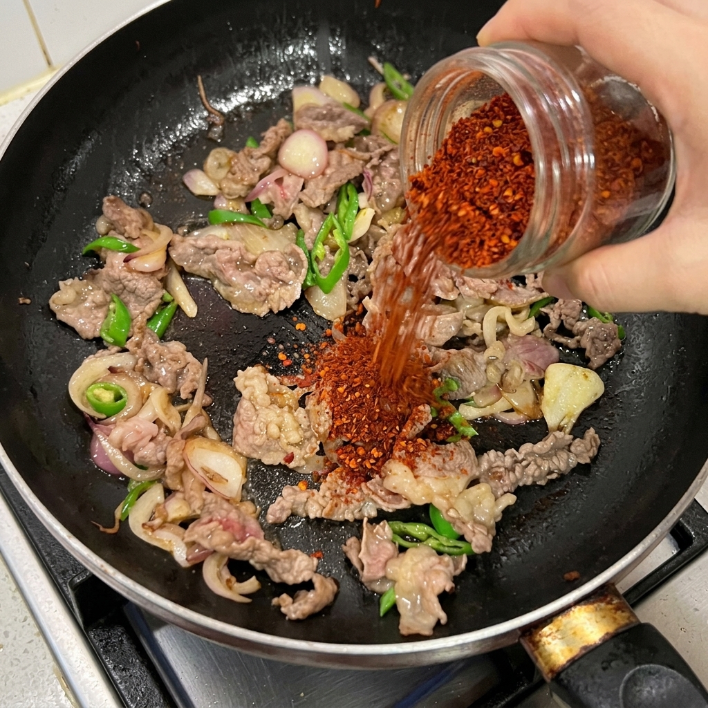 Chile rojo en polvo vertiéndose desde un frasco de vidrio sobre la carne, las cebollas y los chiles verdes en una sartén negra.